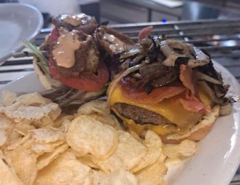 open-face cheeseburger with bacon and grilled veggies on top, served with a side of chips